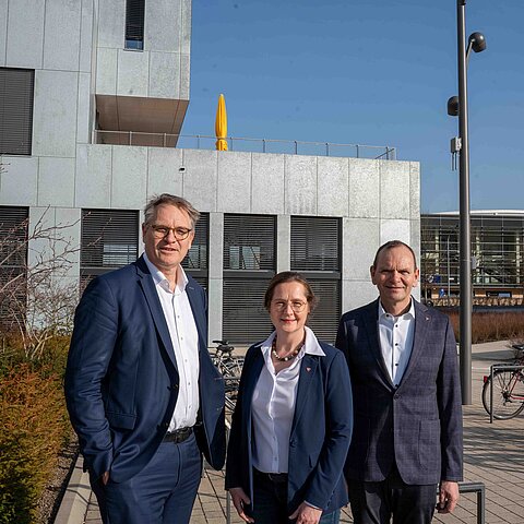 Three people in business attire are standing in sunny weather in front of a modern university building on the campus of the (TH) OWL University of Applied Sciences and Arts, posing for a group photo. Bicycles and other campus buildings can be seen in the background.