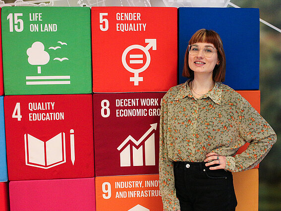 Michelle Buller steht vor den Würfeln der Nachhaltigkeitsziele 4 „Quality Education“, 5 „Gender Equality“, 8 „Decent Work and Economic Growth“ und 15 „Life on Land“ im CIIT der TH OWL. Sie trägt eine beigefarbene geblümte Bluse, schwarze Jeans und eine Brille, stützt ihre linke Hand in die Hüfte und lächelt in die Kamera.