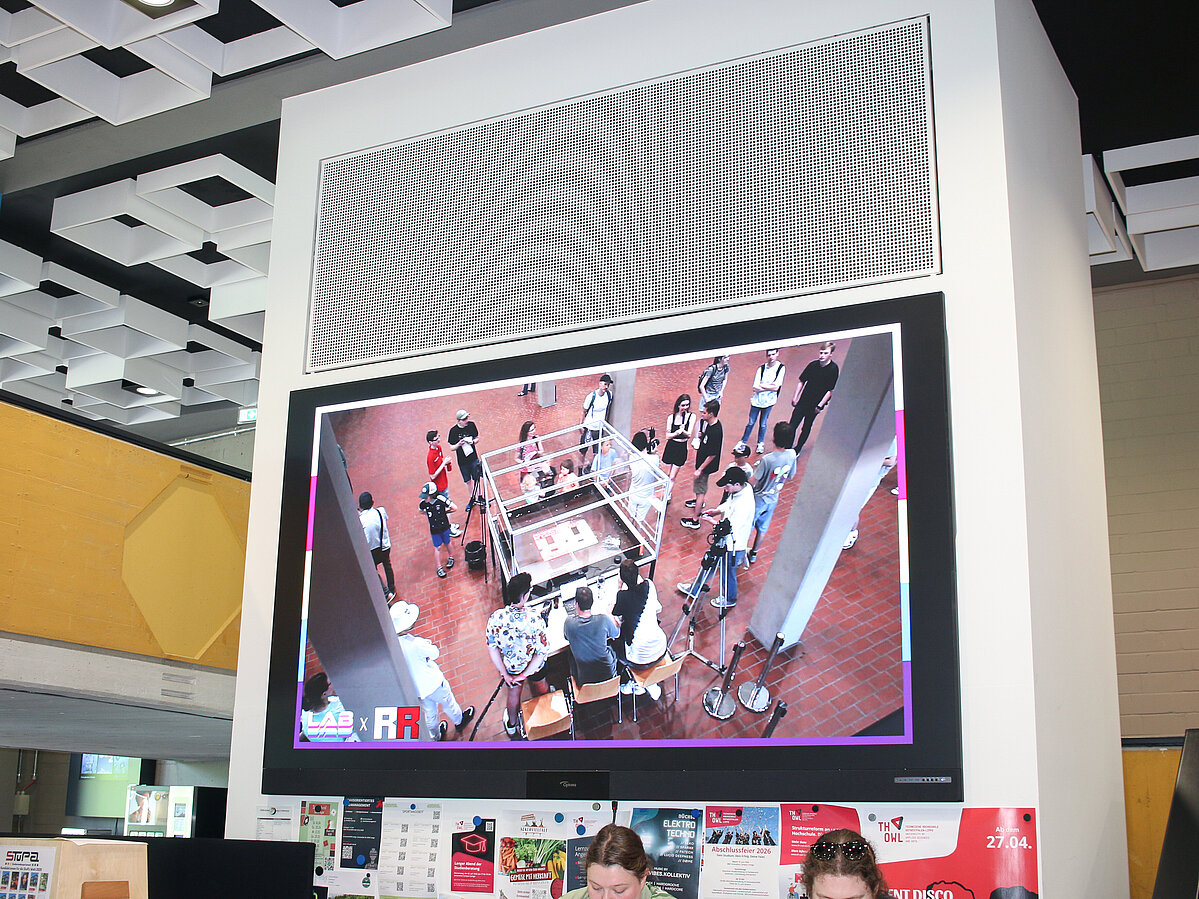A monitor in the lobby of the main building at TH OWL shows a bird's-eye view of the arena. A camera crew is filming the competition and broadcasting the footage.