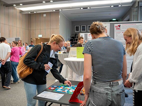 Menschen stehen in einer Ausstellung oder Messehalle an verschiedenen Informationsständen. Im Vordergrund spielen zwei Frauen ein Brettspiel an einem Tisch, während andere Besucherinnen und Besucher Poster und Infotafeln betrachten. Die Atmosphäre wirkt lebendig und interessiert.