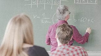 Drei Personen mit dem Rücken zur Kamera, eine Frau und ein Mann sitzen vor einer Tafel, ein weiterer Mann steht an der Tafel und schreibt darauf. Drei Personen mit dem Rücken zur Kamera, eine Frau und ein Mann sitzen vor einer Tafel, ein weiterer Mann steht an der Tafel und schreibt darauf.