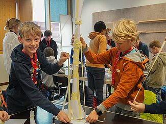 Zwei Jungs zeigen einen Turm aus Papier und Kreppband in die Kamera. In Hintergrund bauen sieben weitere Jungs an ihren Türmen.