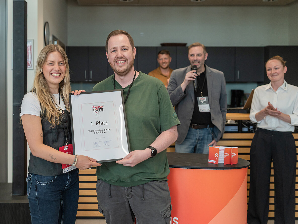 Nächster Meilenstein für Food-Start-up der TH OWL Foto: Eine Frau (links) und ein Mann halten gemeinsam eine gerahmte Urkunde ins Bild. Im Hintergrund stehen zwei Männer und eine Frau und applaudieren.