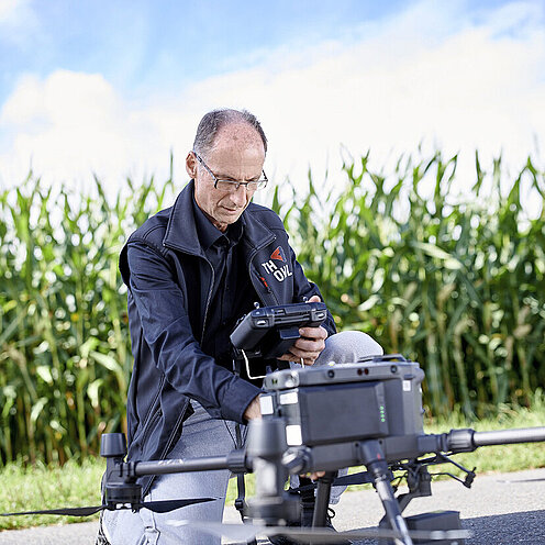 Eine landwirtschaftliche Drohne vor einem Maisfeld. Prof. Dr. Wrenger mit Steuerungsgerät in der Hand kniet daneben.