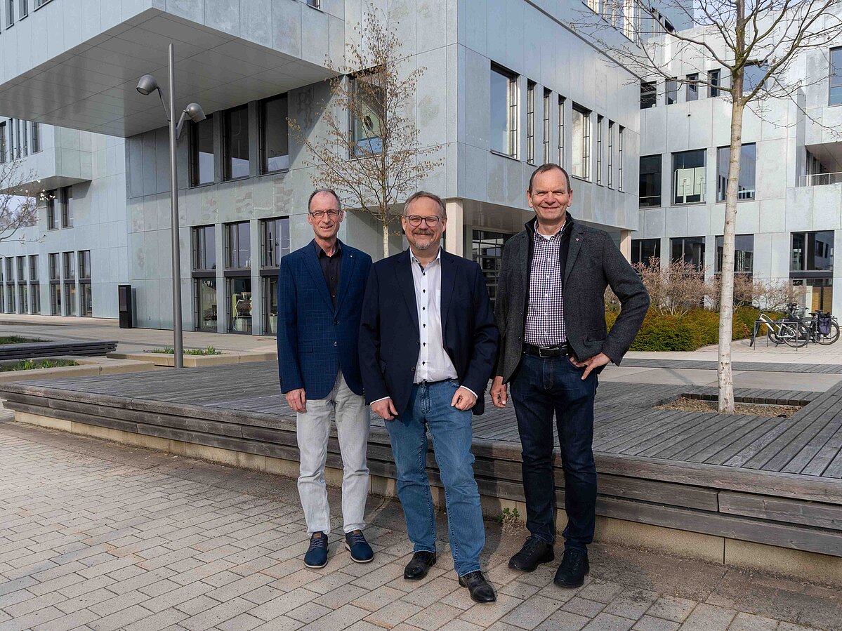 Foto: Drei Männer stehen nebeneinander und blicken in die Kamera. Im Hintergrund ist der InnovationSPIN auf dem Innovation Campus Lemgo zu sehen.