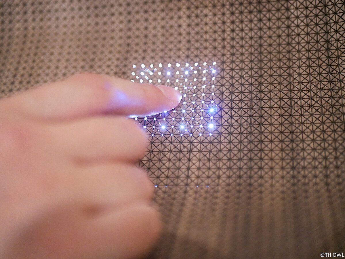 Foto: In einem Panel mit Lochmuster ist ein leuchtendes Touchpad eingelassen. Zu sehen ist eine Hand, die den Knopf drückt.