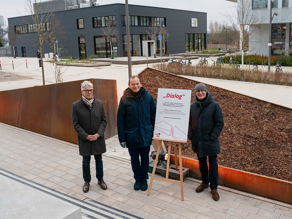 Drei Männer neben einem Schild mit Aufschrift "Dialog" vor einem Kunstwerk