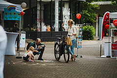 Kind malt mit Kreide auf Boden, Mann hockt daneben, daneben eine Frau mit Fahrrad.