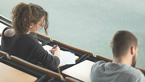 Zwei Studierende sitzen mit dem Rücken zur Kamera in einem Hörsaal und arbeiten