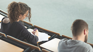 Zwei Studierende sitzen mit dem Rücken zur Kamera in einem Hörsaal und arbeiten
