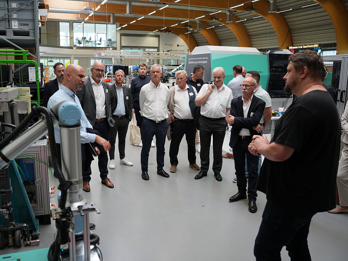 Die Delegation steht in der SmartFactoryOWL. Im Vordergrund spricht Landrat Ali Doğan mit Transfermanager Patrick Spanier und stellt ihm eine Frage. Die übrigen Teilnehmenden verfolgen das Gespräch.