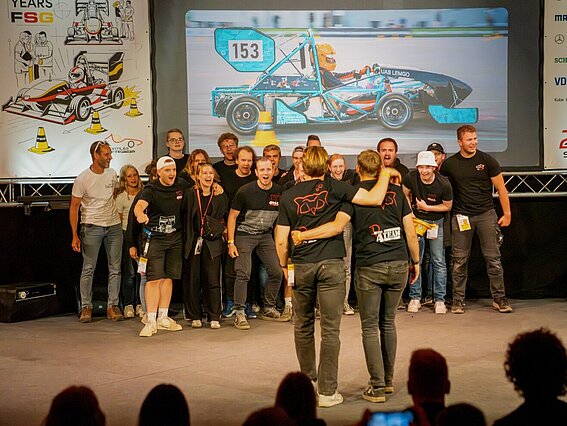Eine große Gruppe Studierender des Racing-Teams der TH OWL jubelt gemeinsam auf einer Bühne. Im Hintergrund ist das eigene Rennfahrzeug auf einer Leinwand zu sehen, das bei der Formula Student Germany in Hockenheim angetreten ist.