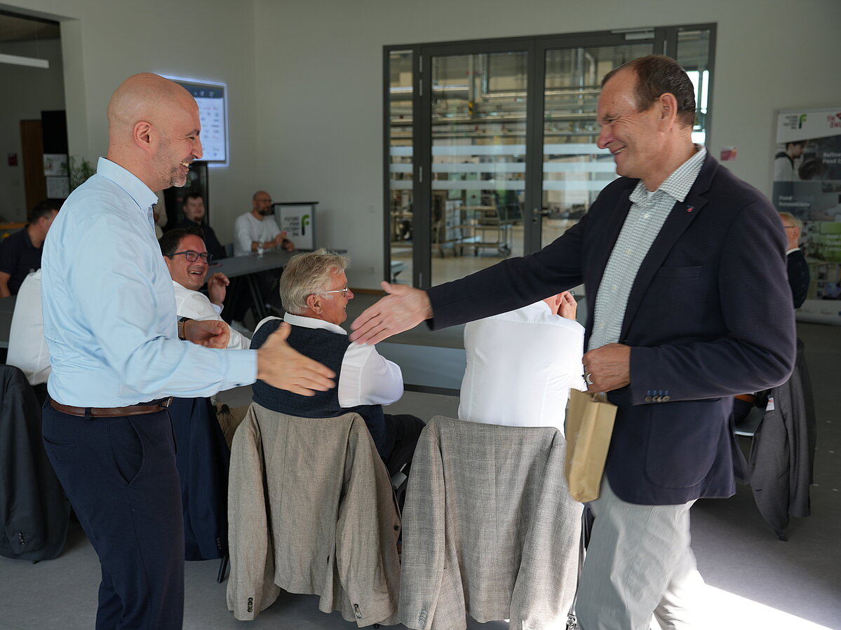  TH-OWL-Präsident Prof. Dr. Jürgen Krahl (rechts) und Landrat Ali Doğan (links) reichen sich die Hand. Doğan überreicht Krahl ein Geschenk aus dem Kreis Minden-Lübbecke. Im Hintergrund sitzt die Delegation an einem Konferenztisch und verfolgt die Szene.