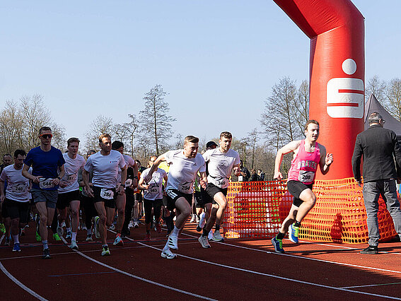 Several runners start at the same time under a red starting arch on a track. In the foreground, participants in athletic wear are sprinting off, while others follow in the background.