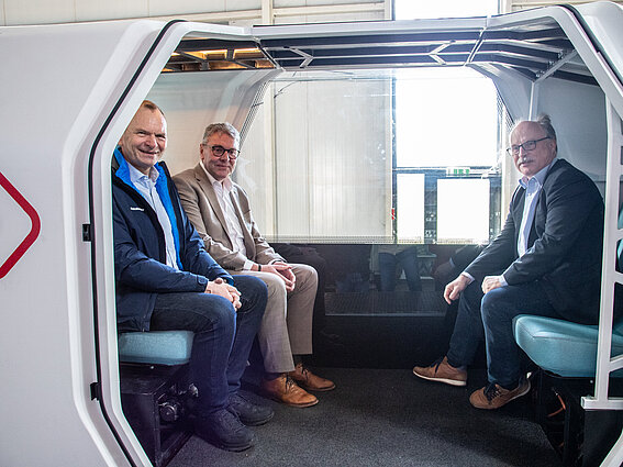 Photo: Three men sit in a MONOCAB test vehicle in an assembly hall. 