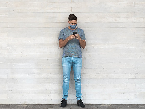 Junger Mann im Grauen Shirt mit Maske und Handy in der Hand