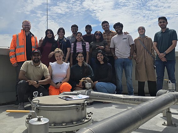 Photo: Eight women and seven men are standing outdoors next to a piece of equipment; pipes are visible in the foreground. 