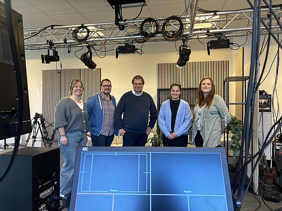 Foto: Drei Frauen und zwei Männer, nebeneinander in einer Reihe stehend,blicken in einem Studio für Bild und Ton lächelnd in die Kamera.
