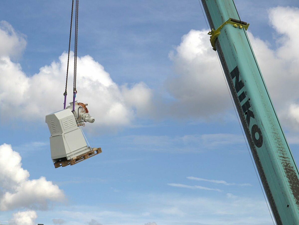 Mischmaschine hängt an Kranarm vor blauem Himmel mit weißen Wölkchen