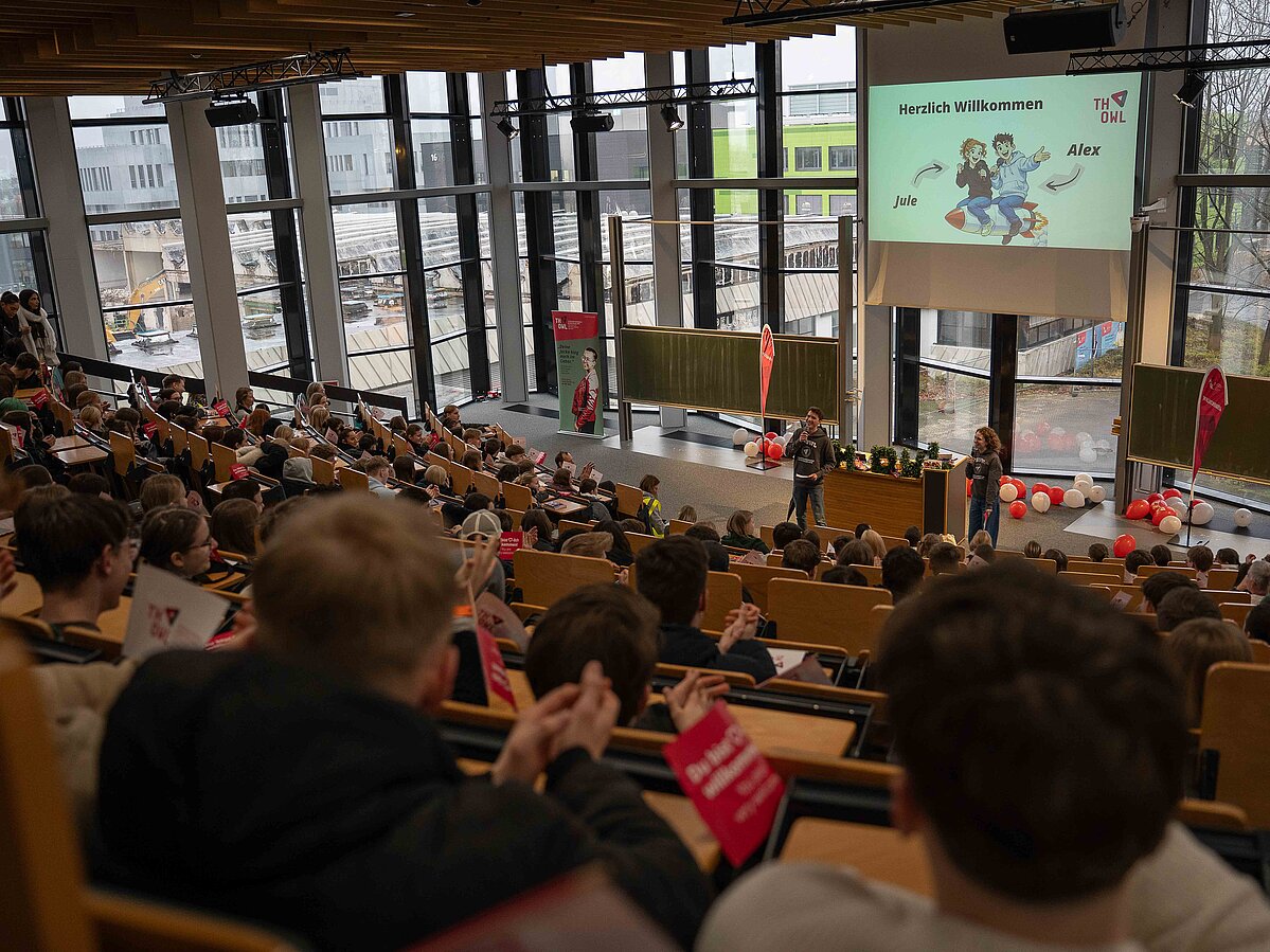 A view into a large lecture hall at TH OWL: numerous students sit in the rows and applaud while two presenters guide the audience through the program on stage. The screen displays the words "Welcome."