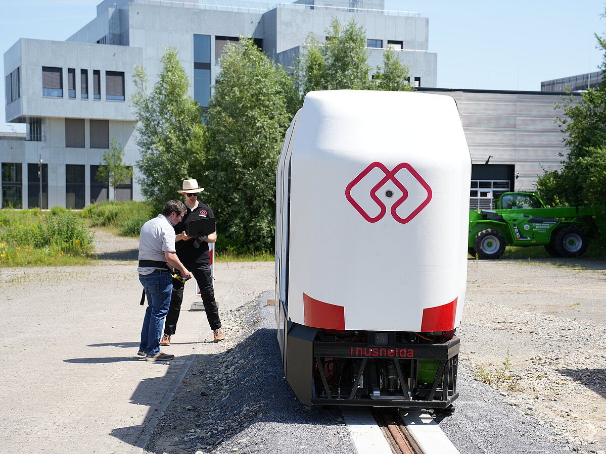 Das MONOCAB fährt auf der Testschiene. Links neben dem Fahrzeug stehen zwei Mitarbeitende des Forschungsteams, die das Fahrzeug fernsteuern.