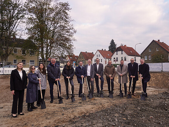 Foto: Eine Gruppe von Menschen stehen mit Schaufeln auf einer Baustelle und setzen zum symbolischen ersten Spatenstich an.