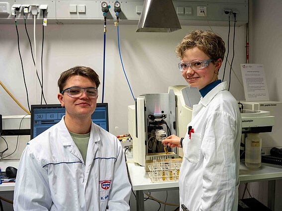 Two trainees in white lab coats and safety goggles are standing at an analytical instrument in a laboratory, conducting a chemical analysis using test tubes.