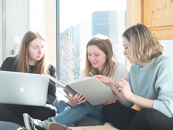 Three young women are sitting together on a sofa, working on their laptops and in their notebooks, and discussing their work in a bright, well-lit study area.