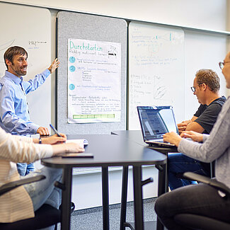 Eine Person zeigt auf ein Flipchart. Eine Personengruppe hört ihm zu.