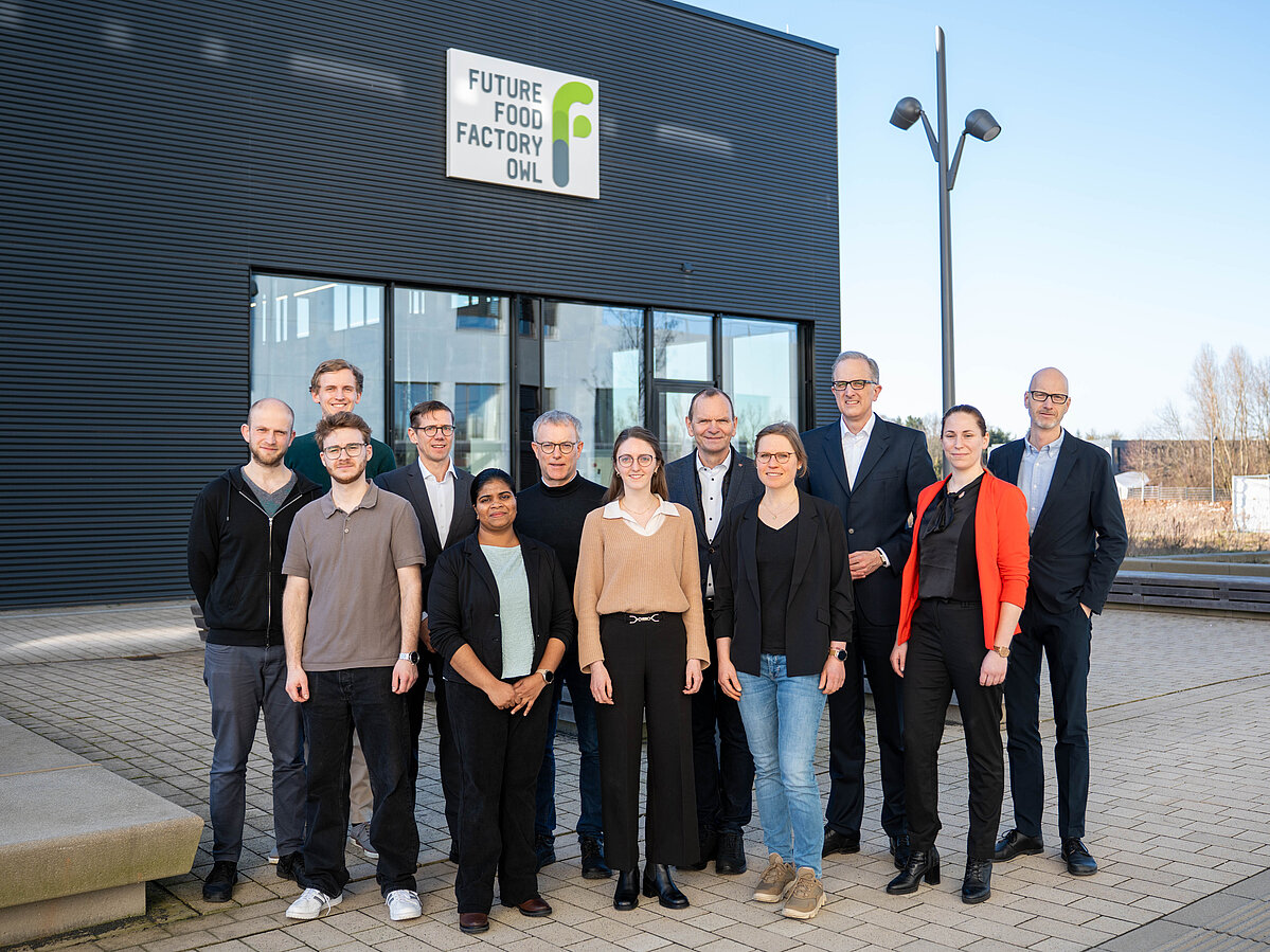Foto: Gruppenbild mit zwölf Personen, im Hintergrund ist eine Außenseite der Future Food Factory in Lemgo zu sehen. 
