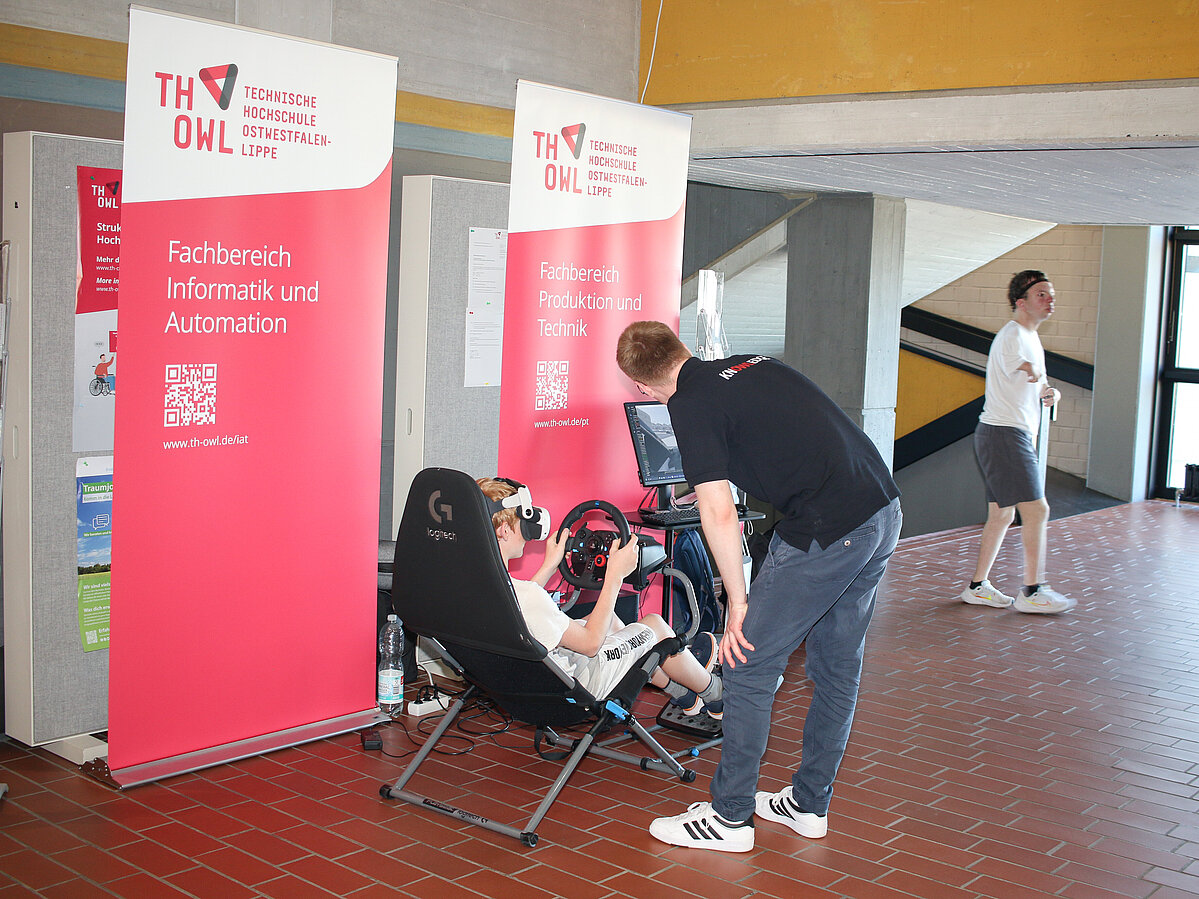 Ein KInd sitzt in einem Fahrsimulator und ein Student betreut die Michmach-Aktion. Er beugt sich zu dem Kind hinunter.
