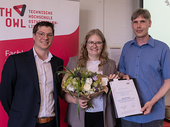 Foto: Eine junge Dame mit einem Blumenstrauß in der Hand wird von zwei Herren links und rechts flankiert. Der Herr rechts hält die Preisurkunde.