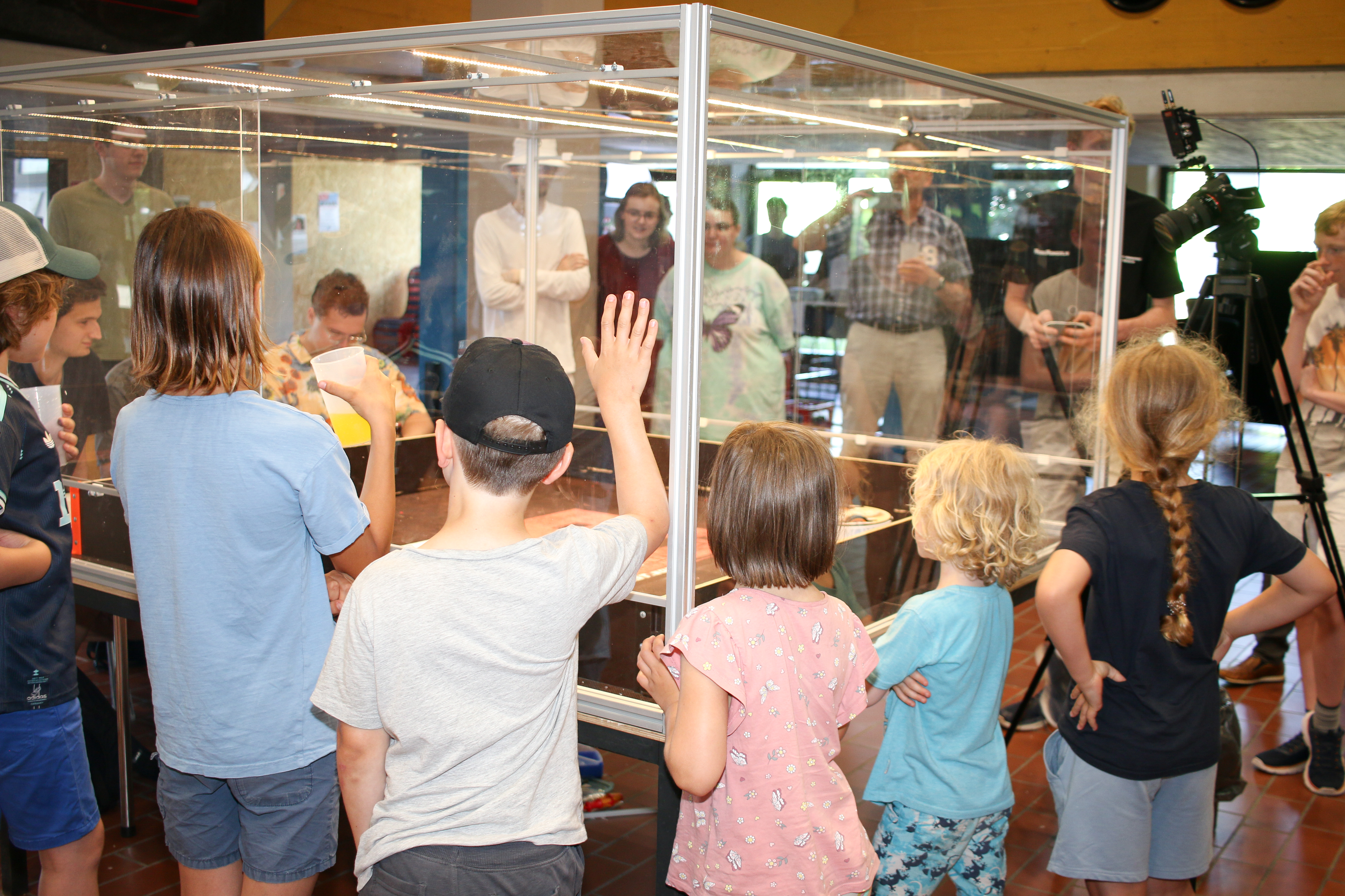 Children are standing at the plexiglass barrier of the arena where robots are fighting each other.