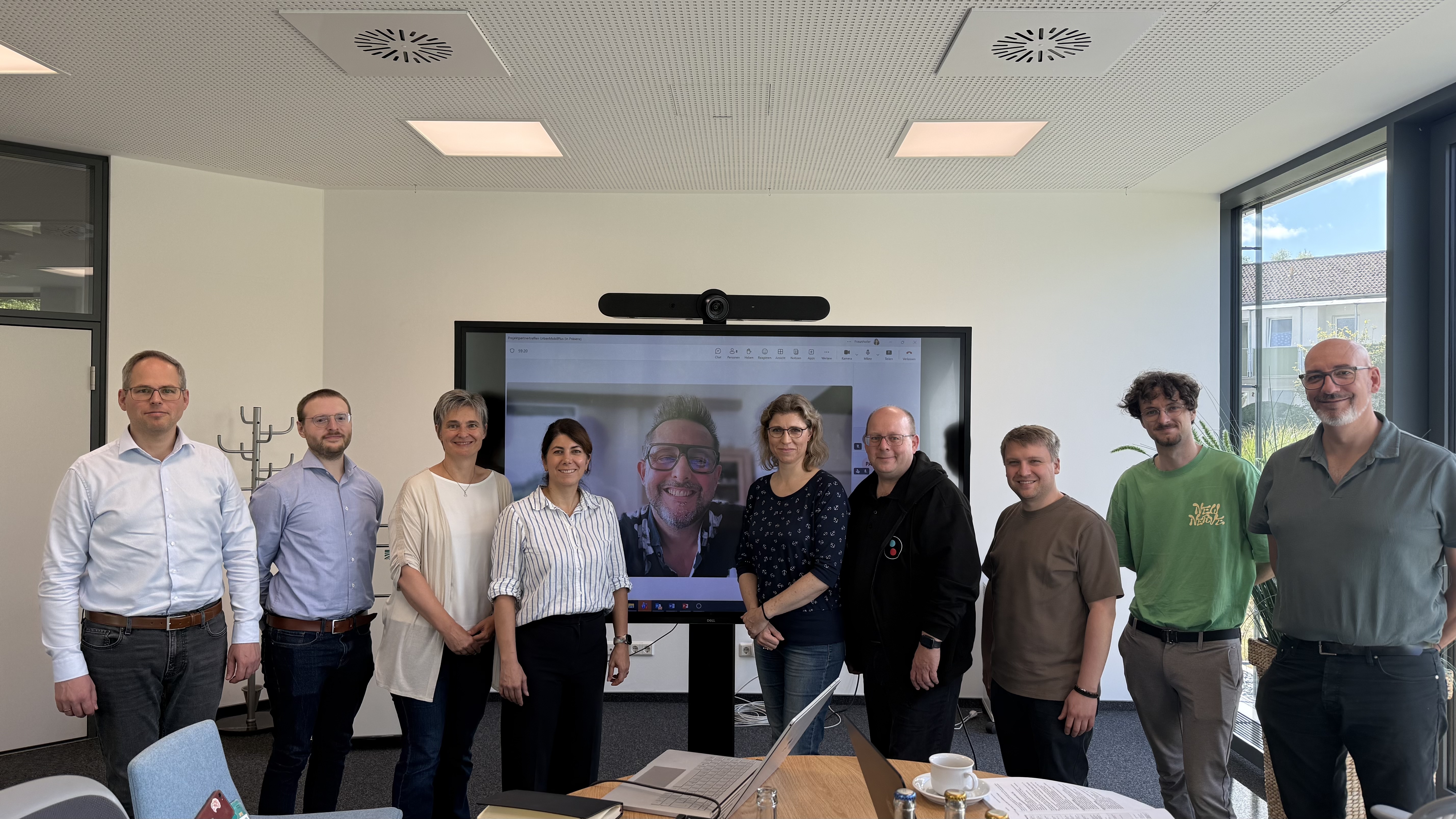 Foto: Zehn Personen, davon drei Damen, sind auf in einem Seminarrraum in einer Reihe nebeneinander zu sehen. Ein Mann ist auf dem großen Monitor dahinter zugeschaltet.